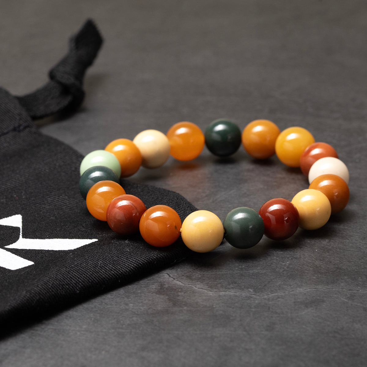 Multicolored beaded bracelet on a dark textured surface with a black bag featuring a white logo.